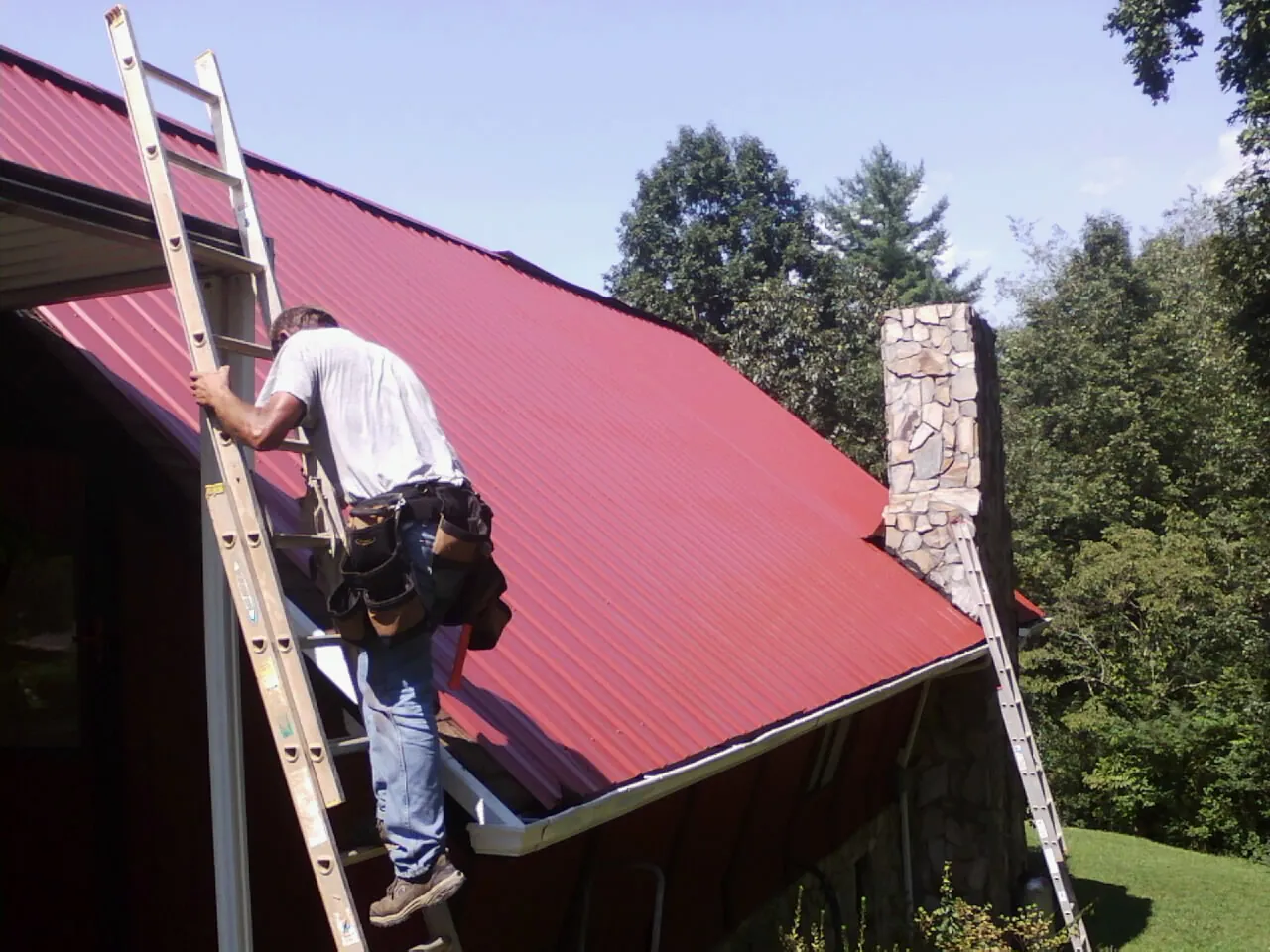 Roofer on a ladder beside a red metal roof and stone chimney — Soffit & Fascia Repair by Roofing Brothers in Lake Milton